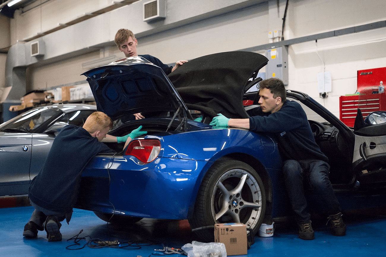 Two men repairing a blue car inside a garage, surrounded by tools and equipment