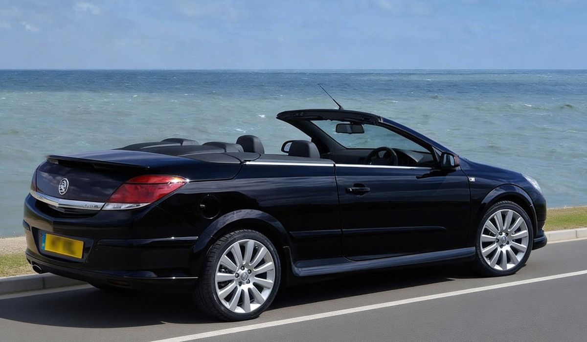 A black convertible car drives along a coastal road with the ocean visible in the background.