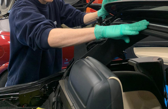 A man in a blue shirt and gloves repairs a car, focused on the engine with tools in hand.